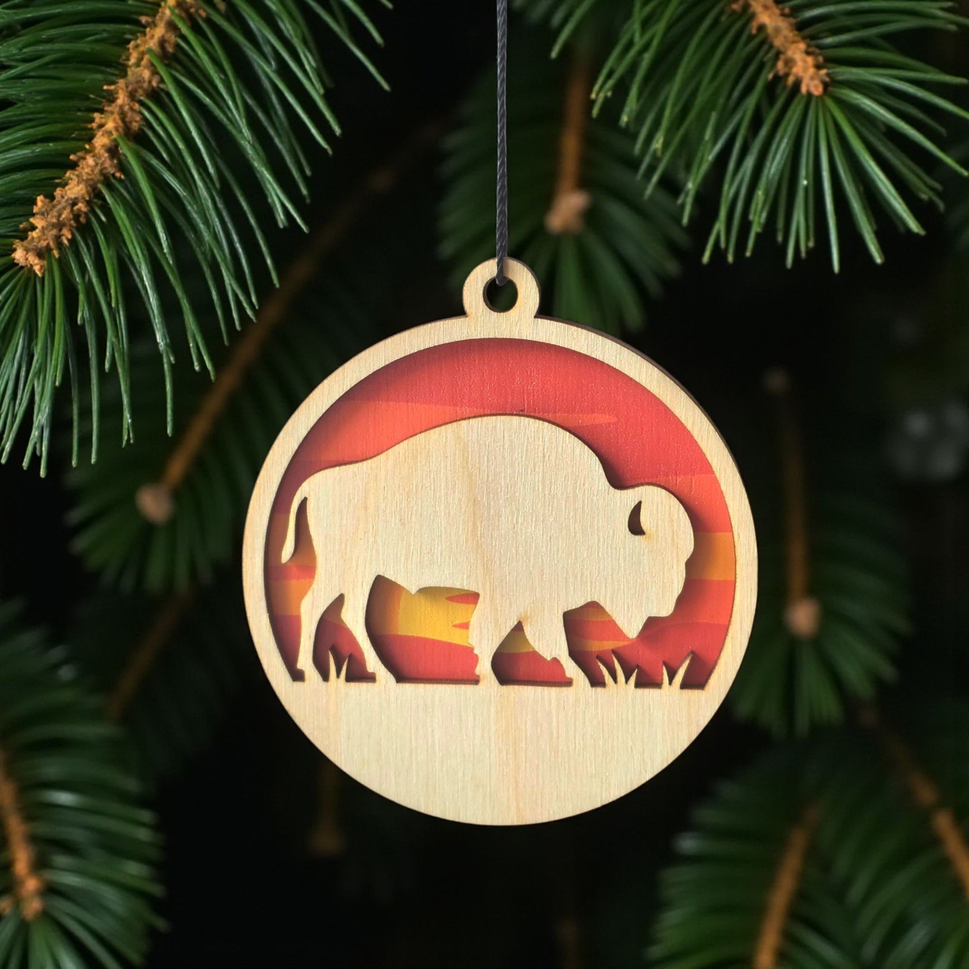 Bison sunset wooden ornament hanging on evergreen tree, layered wood wildlife ornament with warm sunset colors for rustic holiday decor