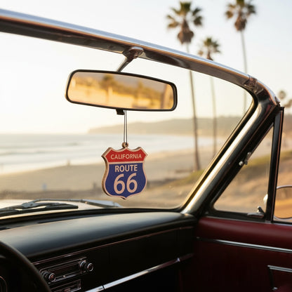 California Route 66 shield ornament hanging from a car rearview mirror beside a coastal highway with palm trees and ocean scenery, vintage Route 66 California travel keepsake