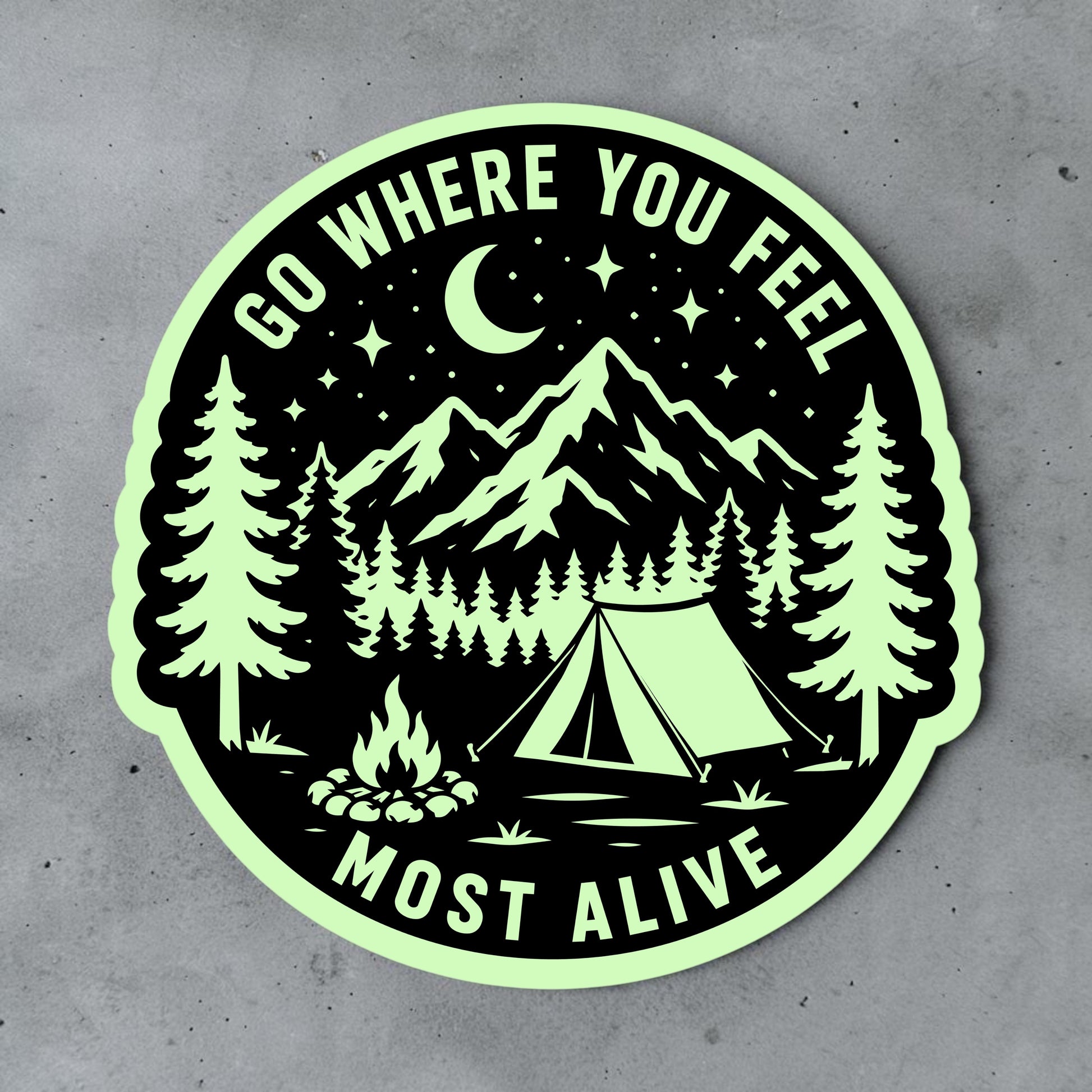 Camping glow in the dark vinyl sticker displayed on a gray surface, featuring mountains, pine trees, campsite, tent, campfire, moon and outdoor adventure quote