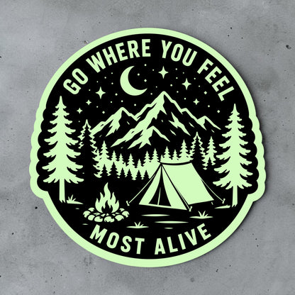 Camping glow in the dark vinyl sticker displayed on a gray surface, featuring mountains, pine trees, campsite, tent, campfire, moon and outdoor adventure quote