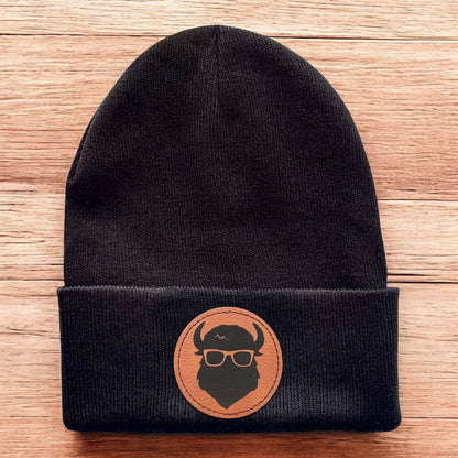 Black Curious Bison knit beanie laid flat on wood background with leatherette logo patch centered on cuff