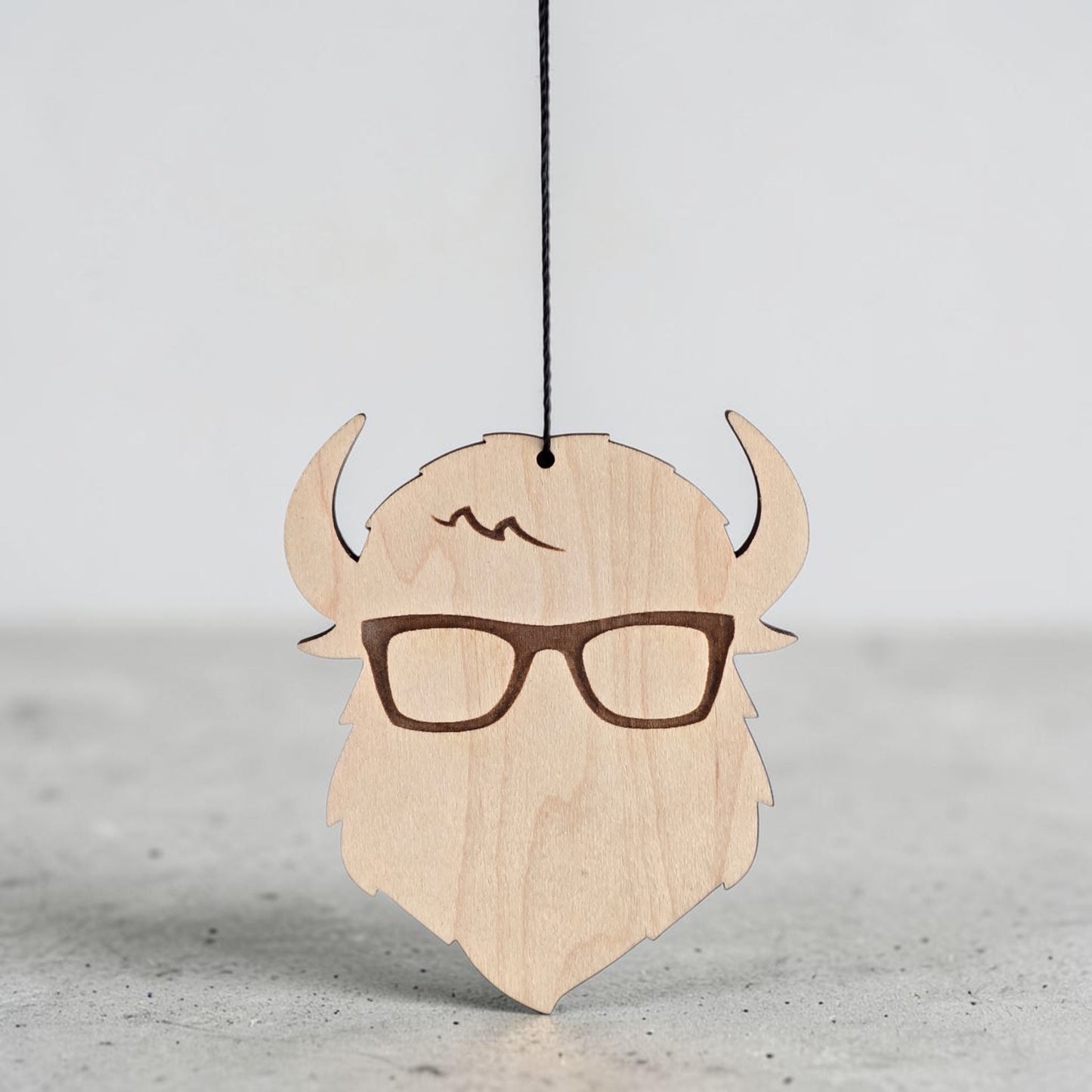 Closeup of Curious Bison wooden ornament hanging by black cord, engraved glasses and logo detail on natural wood decorative ornament