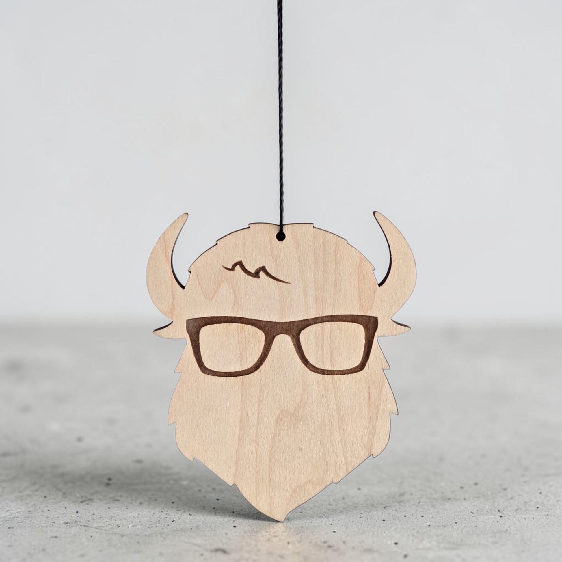 Closeup of Curious Bison wooden ornament hanging by black cord, engraved glasses and logo detail on natural wood decorative ornament