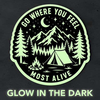 Glow in the dark camping sticker shown glowing pale green against a dark background with mountain, forest, tent and campfire artwork and Go Where You Feel Most Alive text