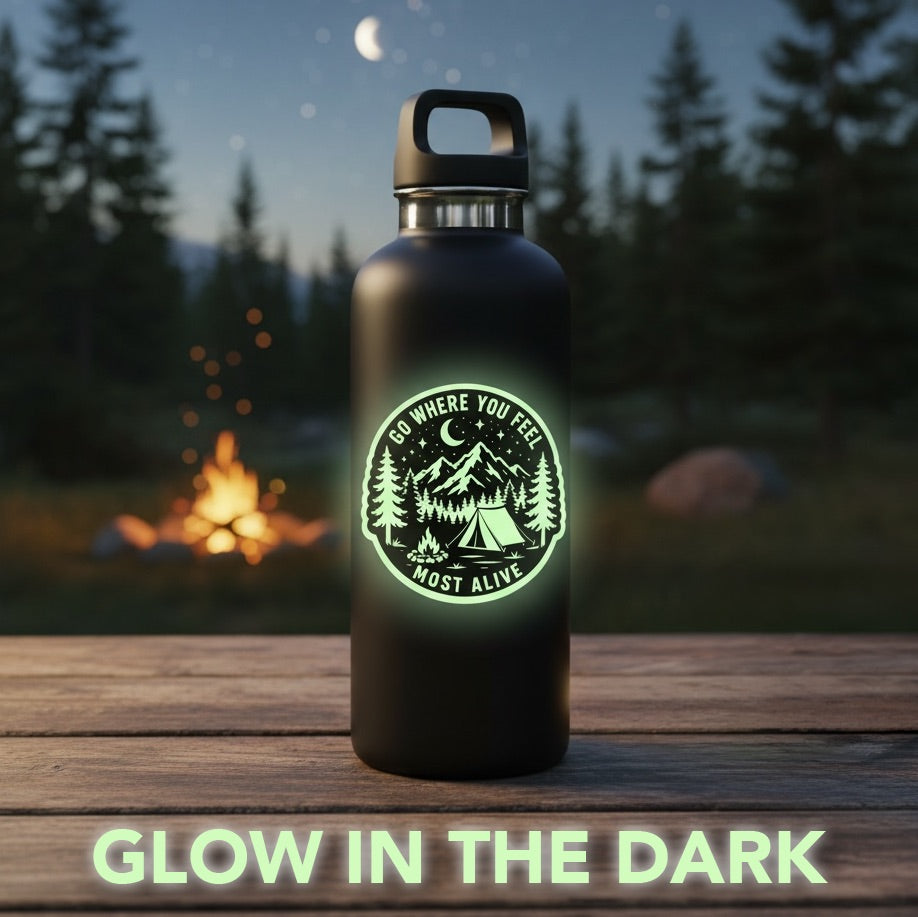 Glow in the dark camping vinyl sticker glowing on a black water bottle at a nighttime campsite with pine trees, crescent moon and campfire in the background