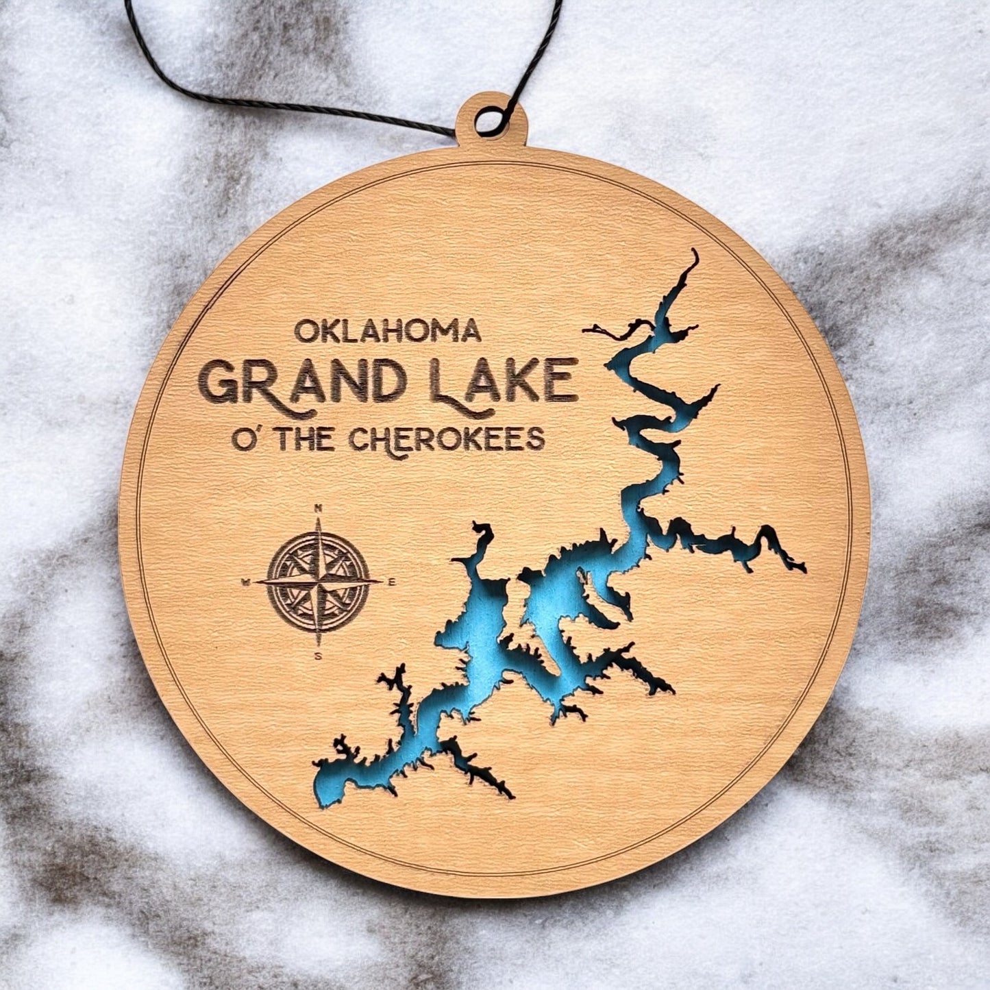 Close up of Grand Lake Oklahoma wooden ornament showing shoreline cutout, engraved text, compass detail, and blue painted water