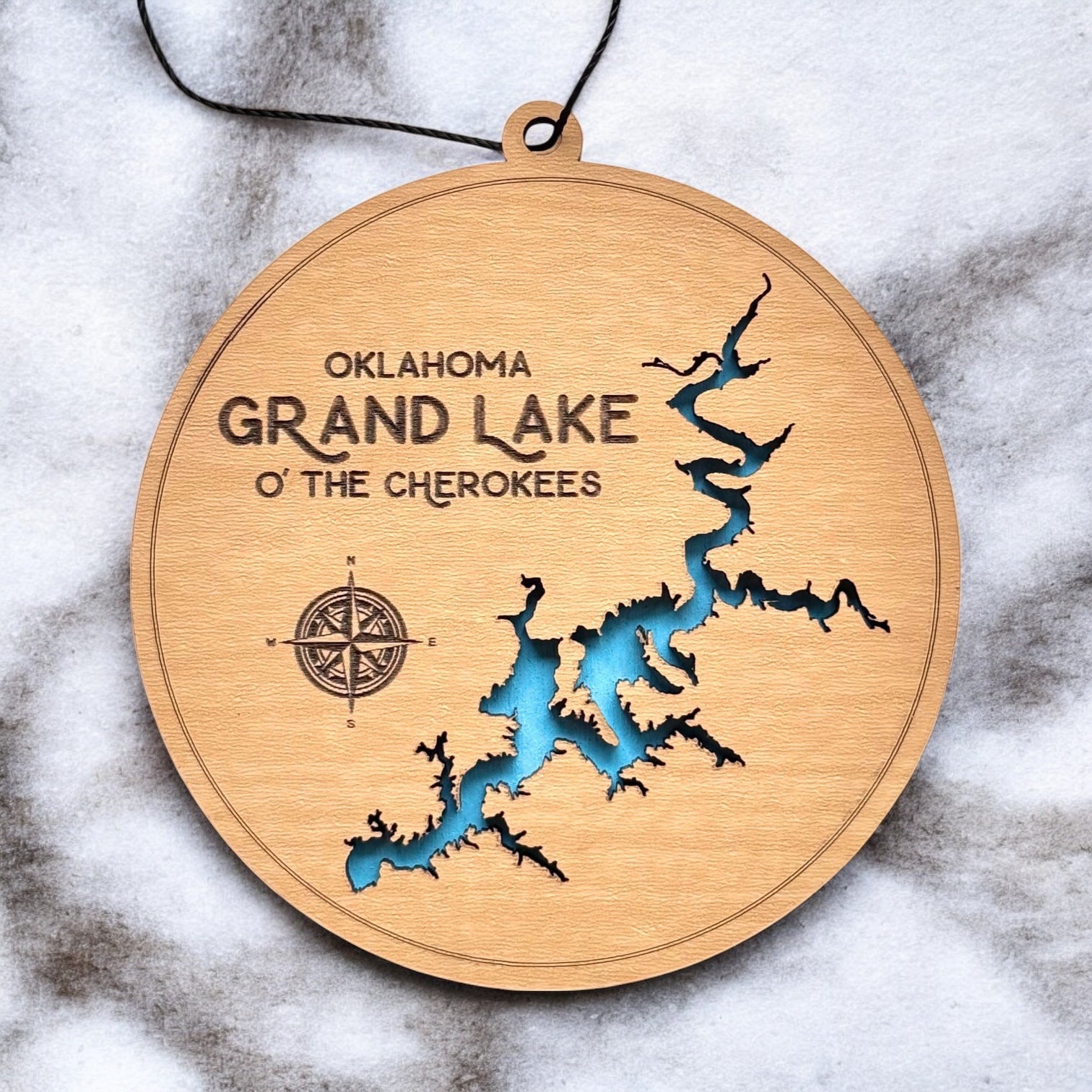 Close up of Grand Lake Oklahoma wooden ornament showing shoreline cutout, engraved text, compass detail, and blue painted water