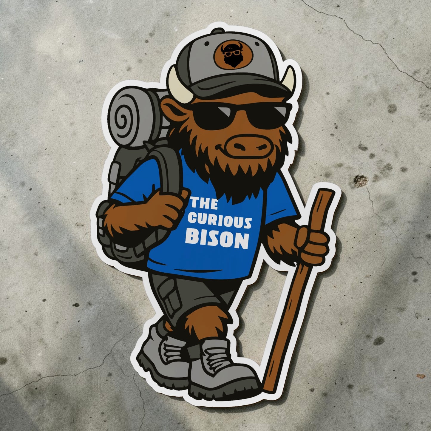 die cut Kerf the Curious Bison hiking mascot sticker on textured concrete surface showing backpack sunglasses walking stick and blue Curious Bison shirt
