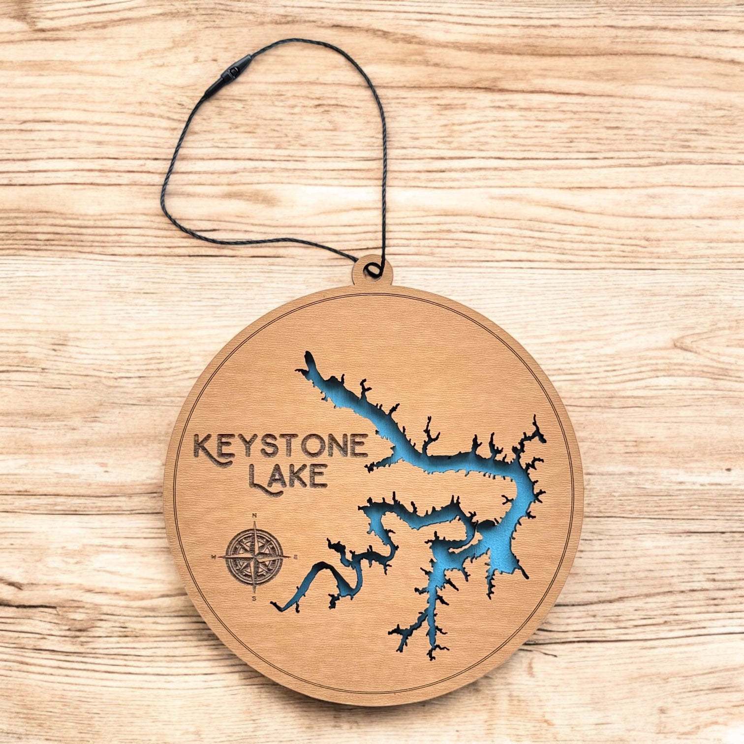 Keystone Lake layered wood ornament shown on wood surface, round lake map ornament with engraved text and blue water detail