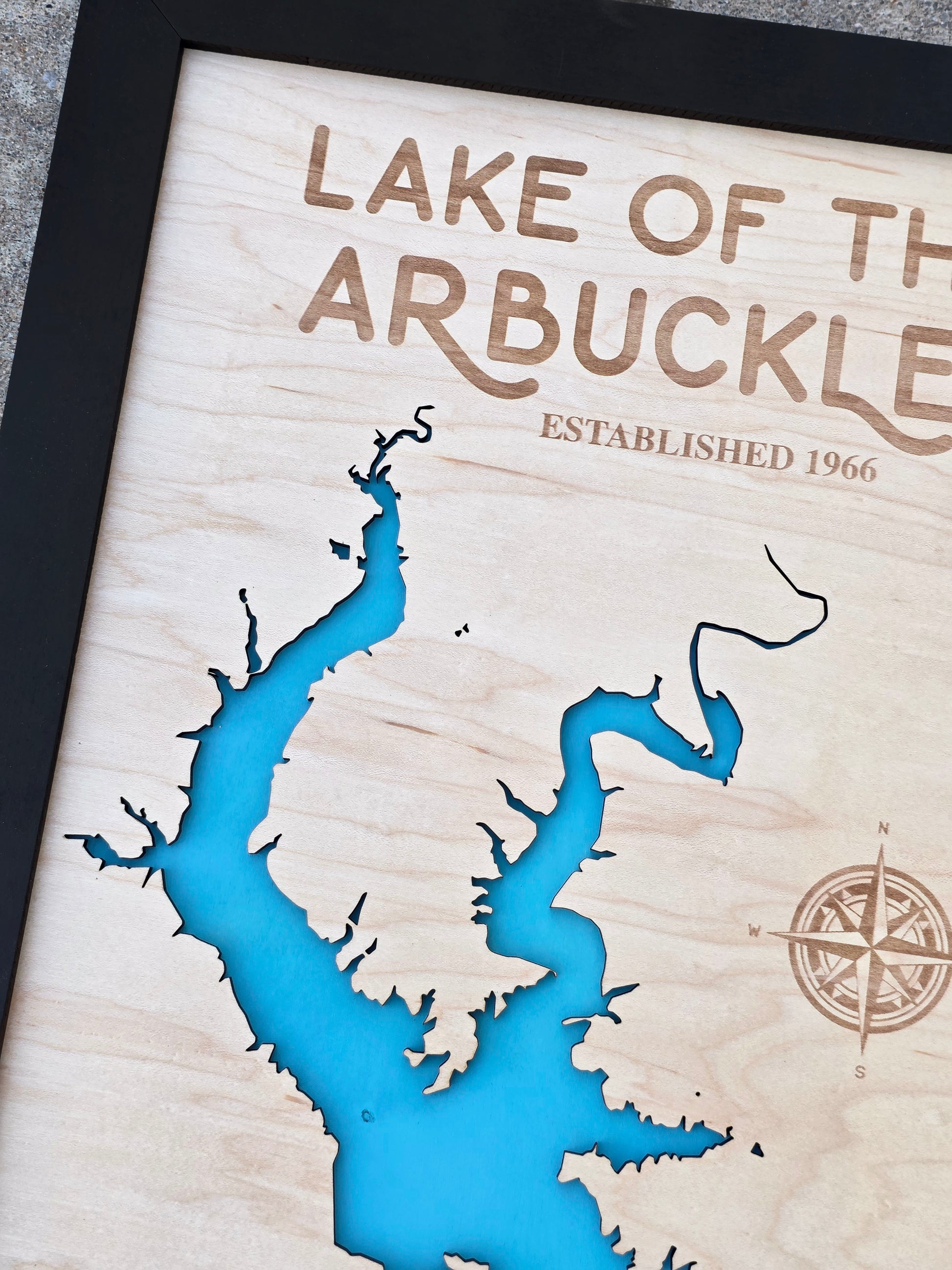 Close up of Lake of the Arbuckles wooden map showing shoreline cutout, blue water layer, engraved compass, and natural wood grain