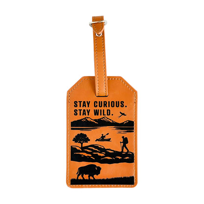 Front view of tan leatherette luggage tag with adjustable buckle strap and laser engraved Stay Curious Stay Wild outdoor scene featuring mountains, water, kayaking, hiking, and a bison