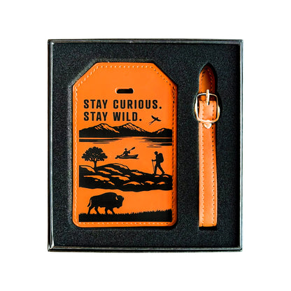 Tan Oklahoma adventure leatherette luggage tag displayed in a black gift box with matching buckle strap, Stay Curious Stay Wild wording, mountains, kayaker, hiker, and bison design for a travel gift or souvenir
