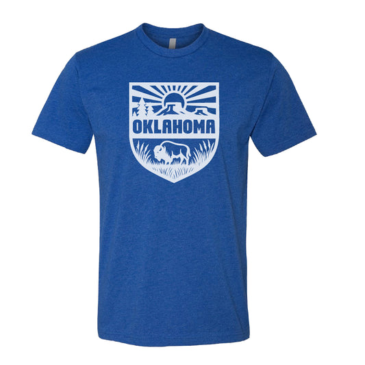 Heather royal blue Oklahoma Adventure t shirt with white shield graphic featuring bison, prairie grass, mesas, trees and sunrise, Oklahoma pride graphic tee