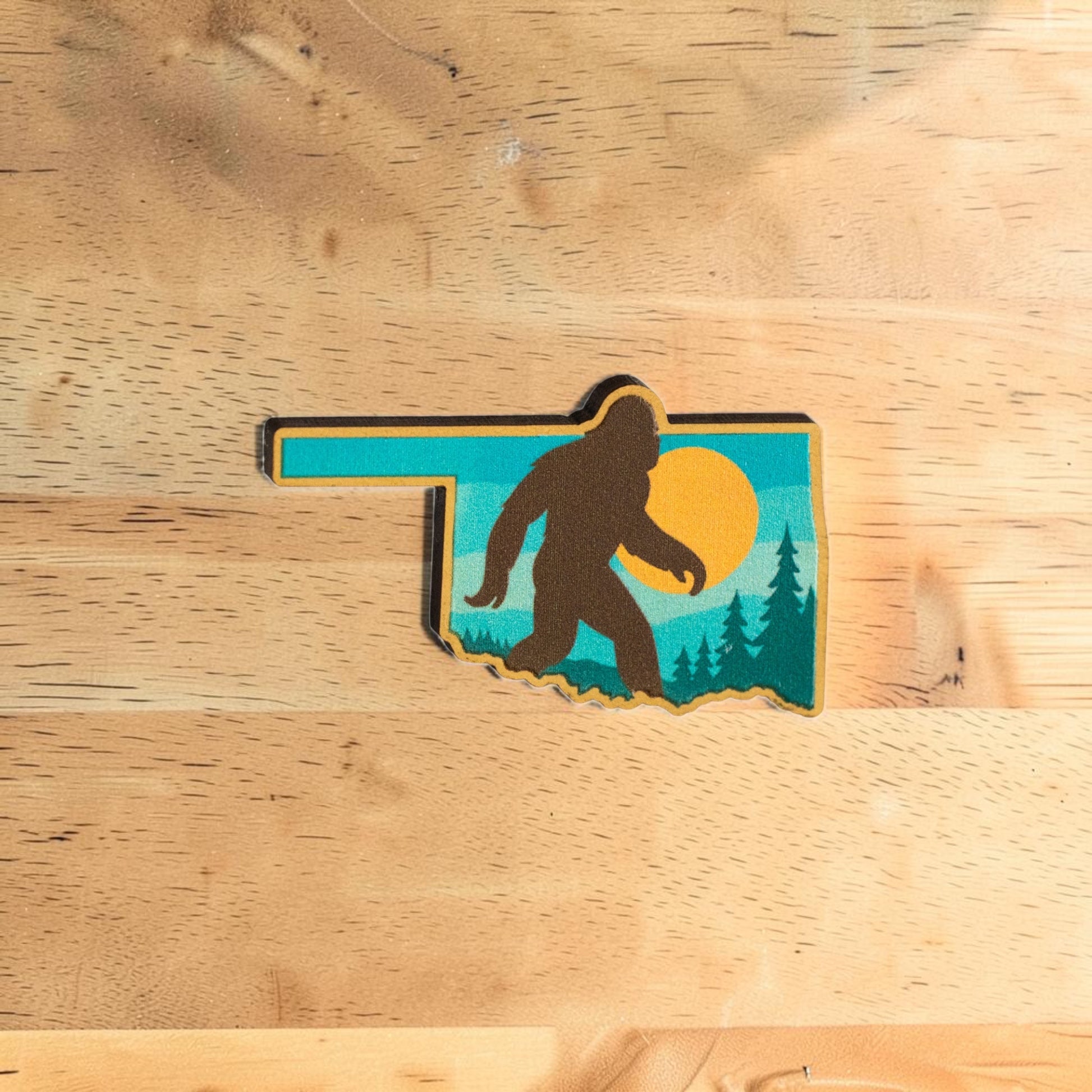 Oklahoma Bigfoot lapel pin made from Eco Wood Birch featuring state shape with walking Sasquatch silhouette, sunset sky, trees, and mountains on wood table