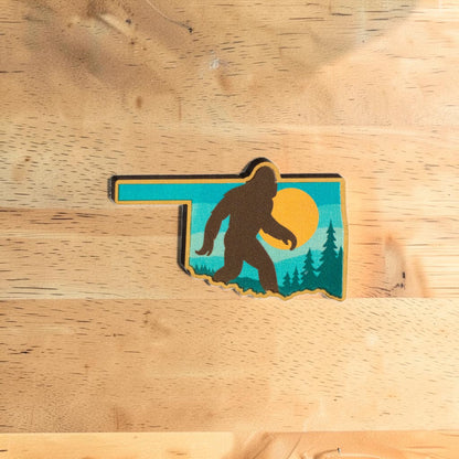 Oklahoma Bigfoot lapel pin made from Eco Wood Birch featuring state shape with walking Sasquatch silhouette, sunset sky, trees, and mountains on wood table