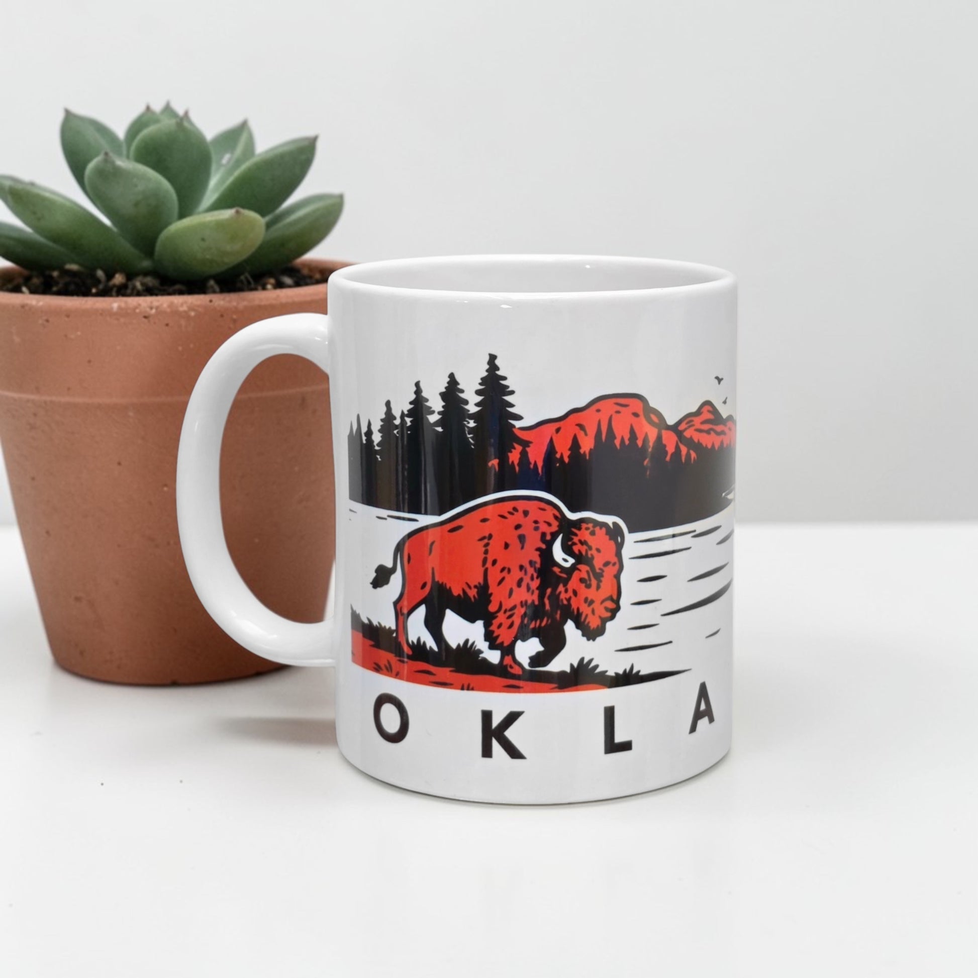 Oklahoma bison sunset 11oz coffee mug showing red sunset mountain landscape with pine trees and American bison walking near water, ceramic Oklahoma wildlife souvenir mug