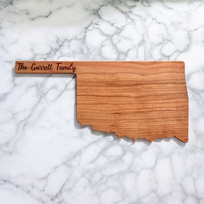 Personalized Oklahoma-shaped cherry wood charcuterie board with custom engraved family name on marble background.