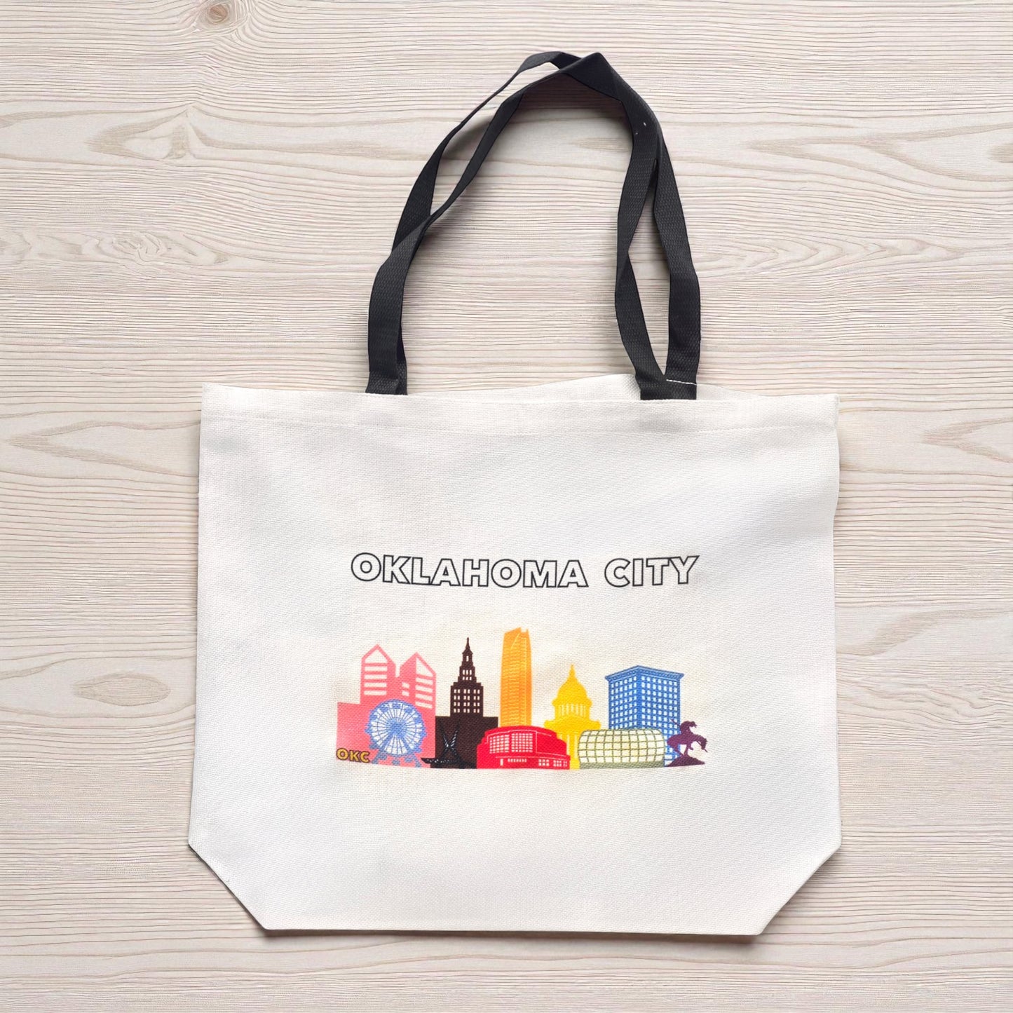 Flat lay of large Oklahoma City pride skyline tote bag on wood surface, reusable pride tote with black handles