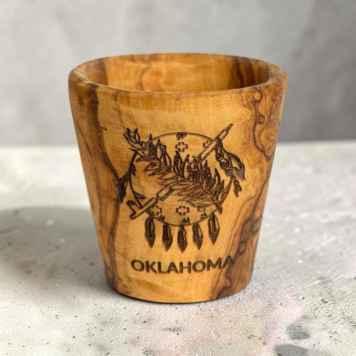 Laser engraved Oklahoma flag olive wood shot glass standing on a tabletop, natural wooden Oklahoma souvenir and state gift with visible wood grain