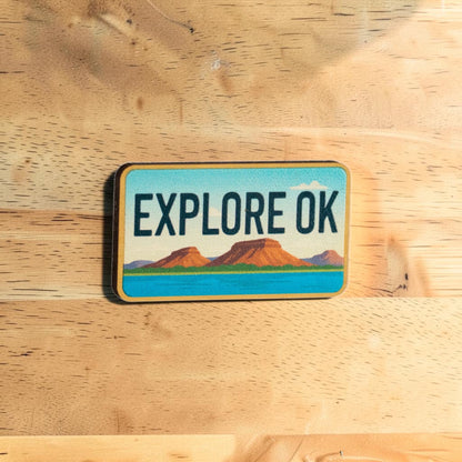 Oklahoma lapel pin with Explore OK text in vintage license plate style featuring scenic mountains and blue water, collectible Oklahoma souvenir pin displayed on wood surface