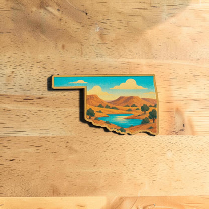 Oklahoma lapel pin made from Eco Wood Birch featuring state shape with scenic blue lake, plateaus, trees, and open sky on wood table