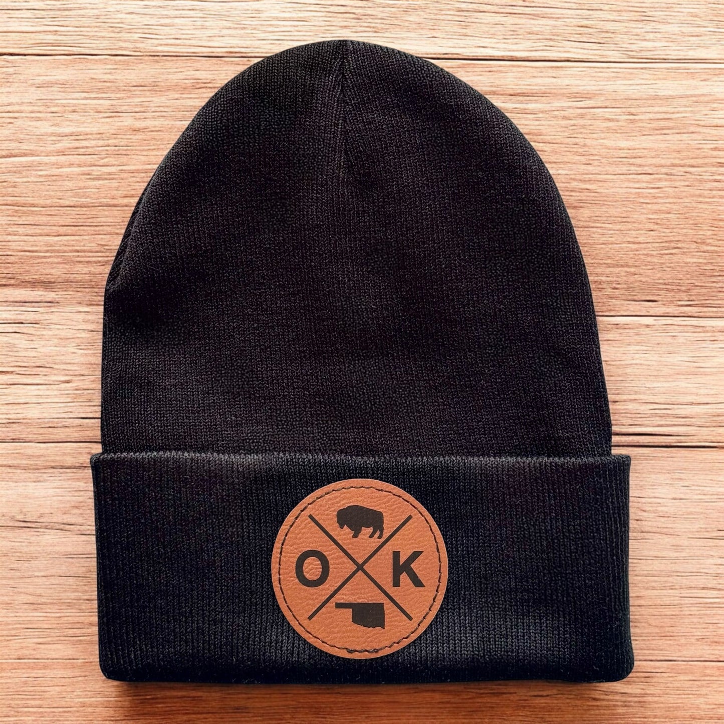 Black Oklahoma OK X beanie displayed on wood background, knit cuffed hat with engraved leatherette patch for Oklahoma souvenir gift