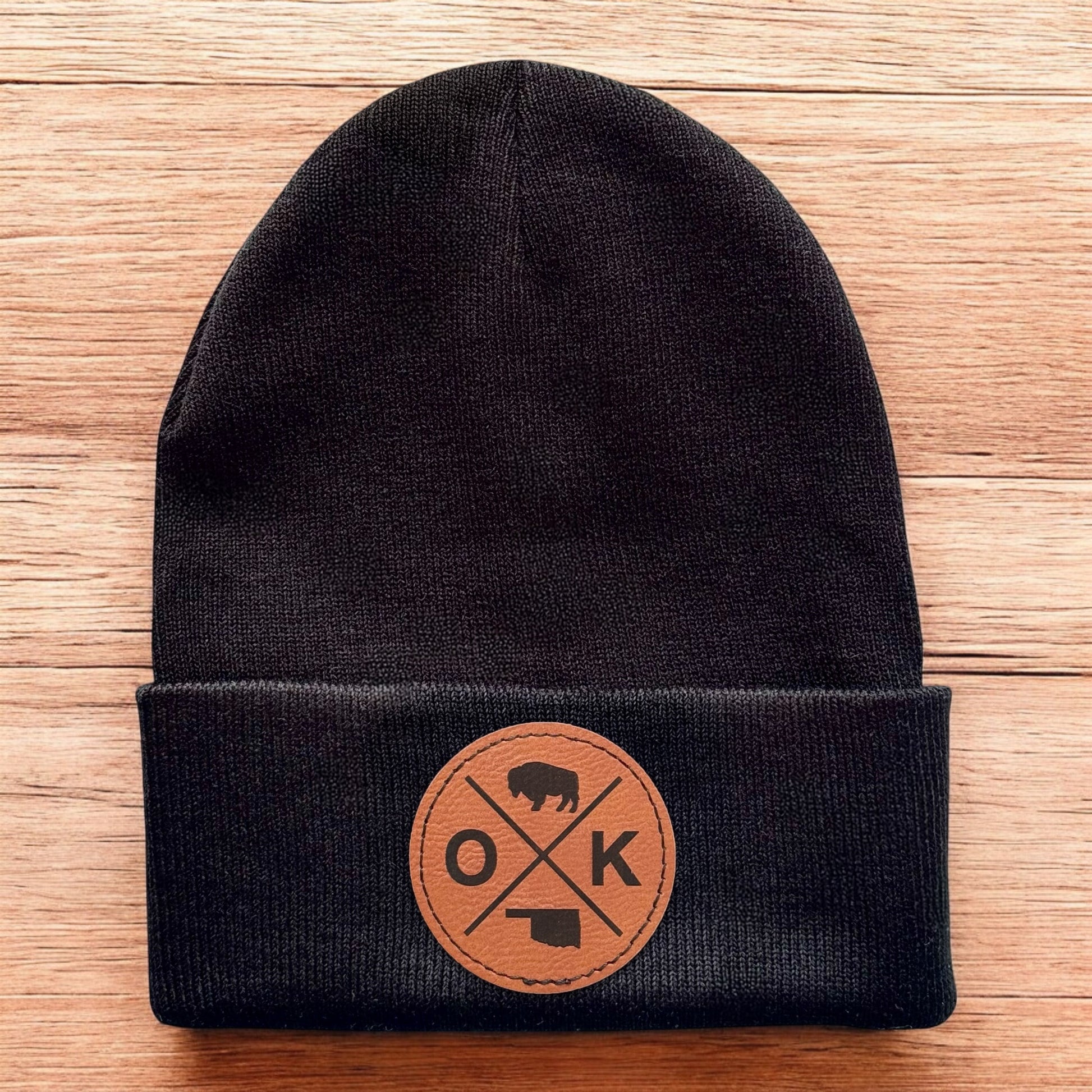Black Oklahoma OK X beanie displayed on wood background, knit cuffed hat with engraved leatherette patch for Oklahoma souvenir gift