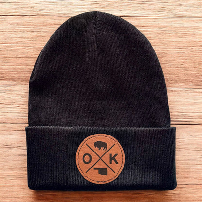 Black Oklahoma OK X beanie displayed on wood background, knit cuffed hat with engraved leatherette patch for Oklahoma souvenir gift