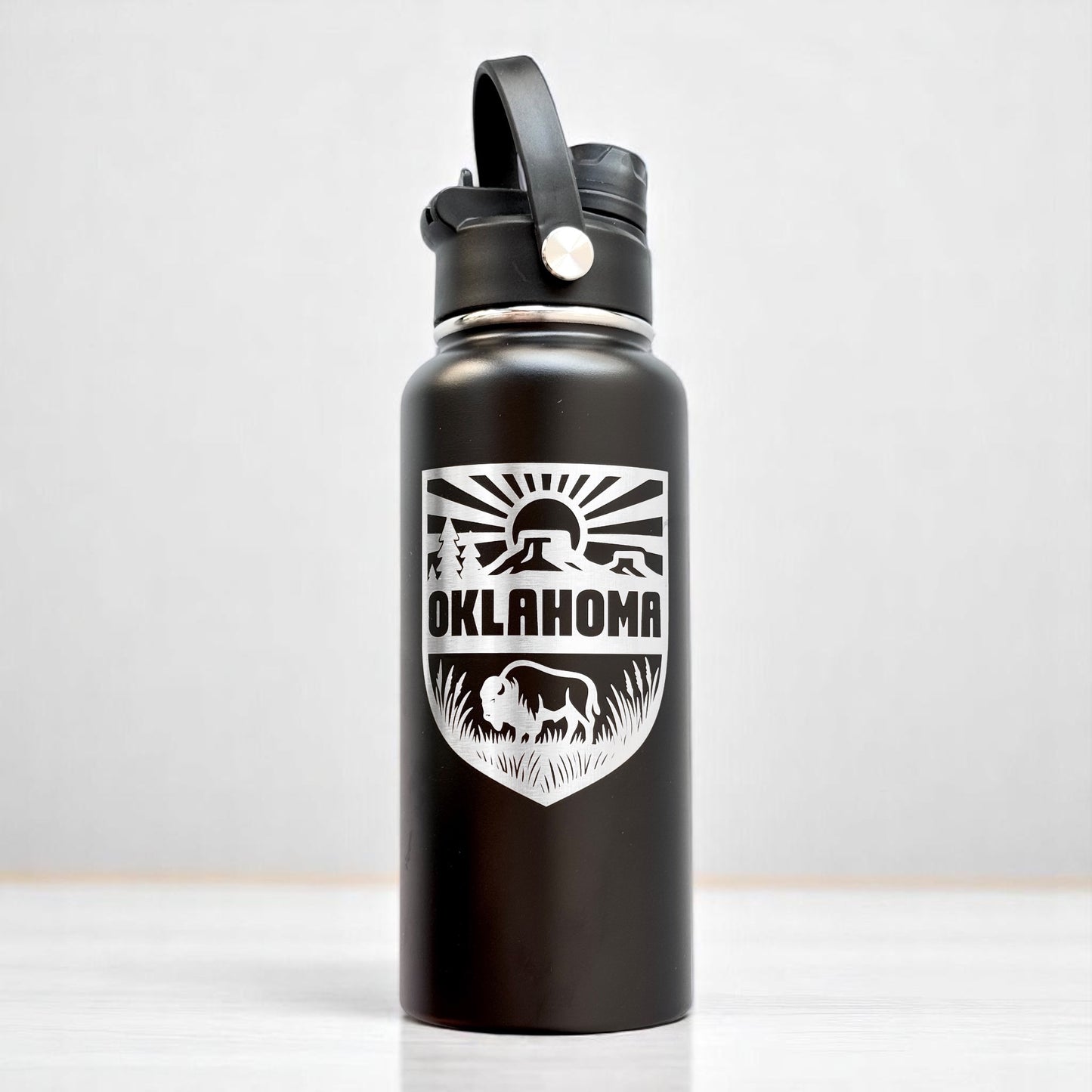 Angled view of black Oklahoma adventure water bottle with laser engraved shield design and dual lid