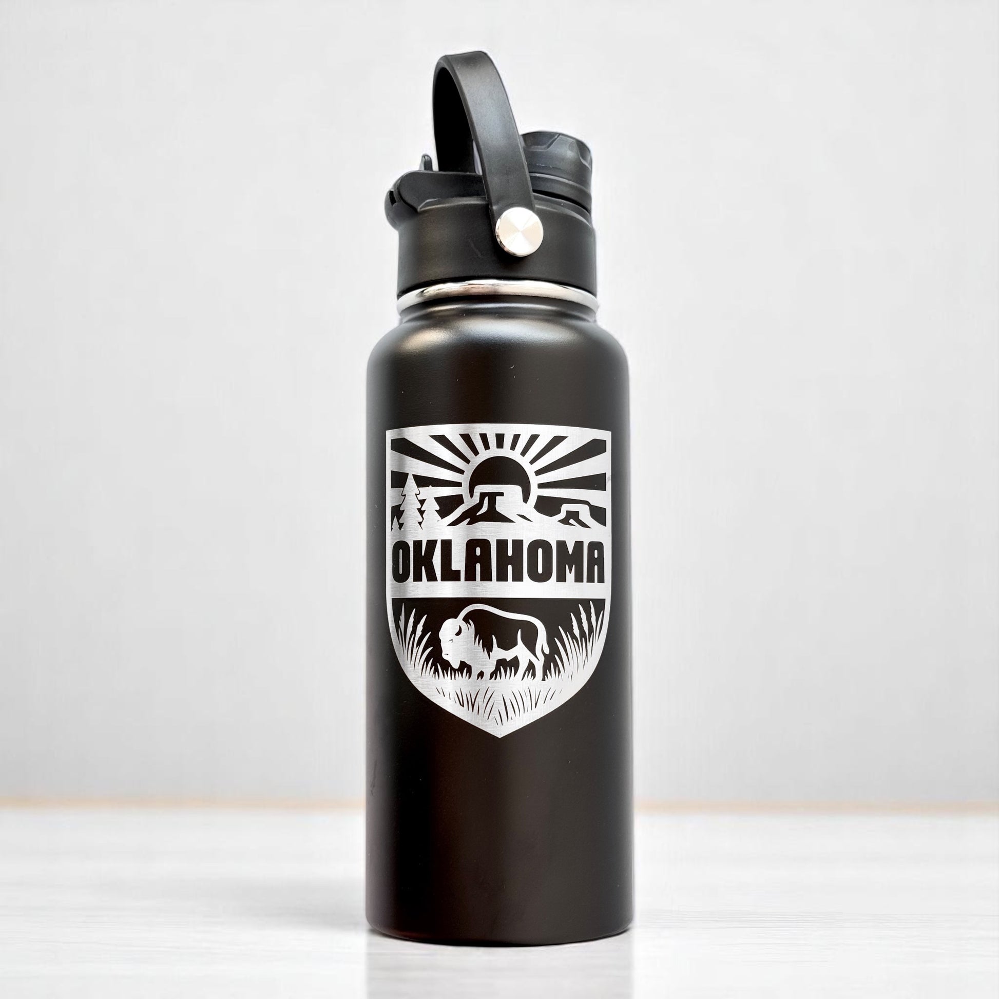 Angled view of black Oklahoma adventure water bottle with laser engraved shield design and dual lid