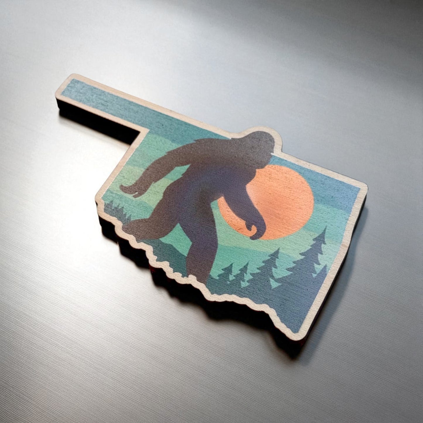 Angled view of Oklahoma Bigfoot Sasquatch wooden magnet showing state outline shape and colorful forest sunset design, novelty Oklahoma magnet