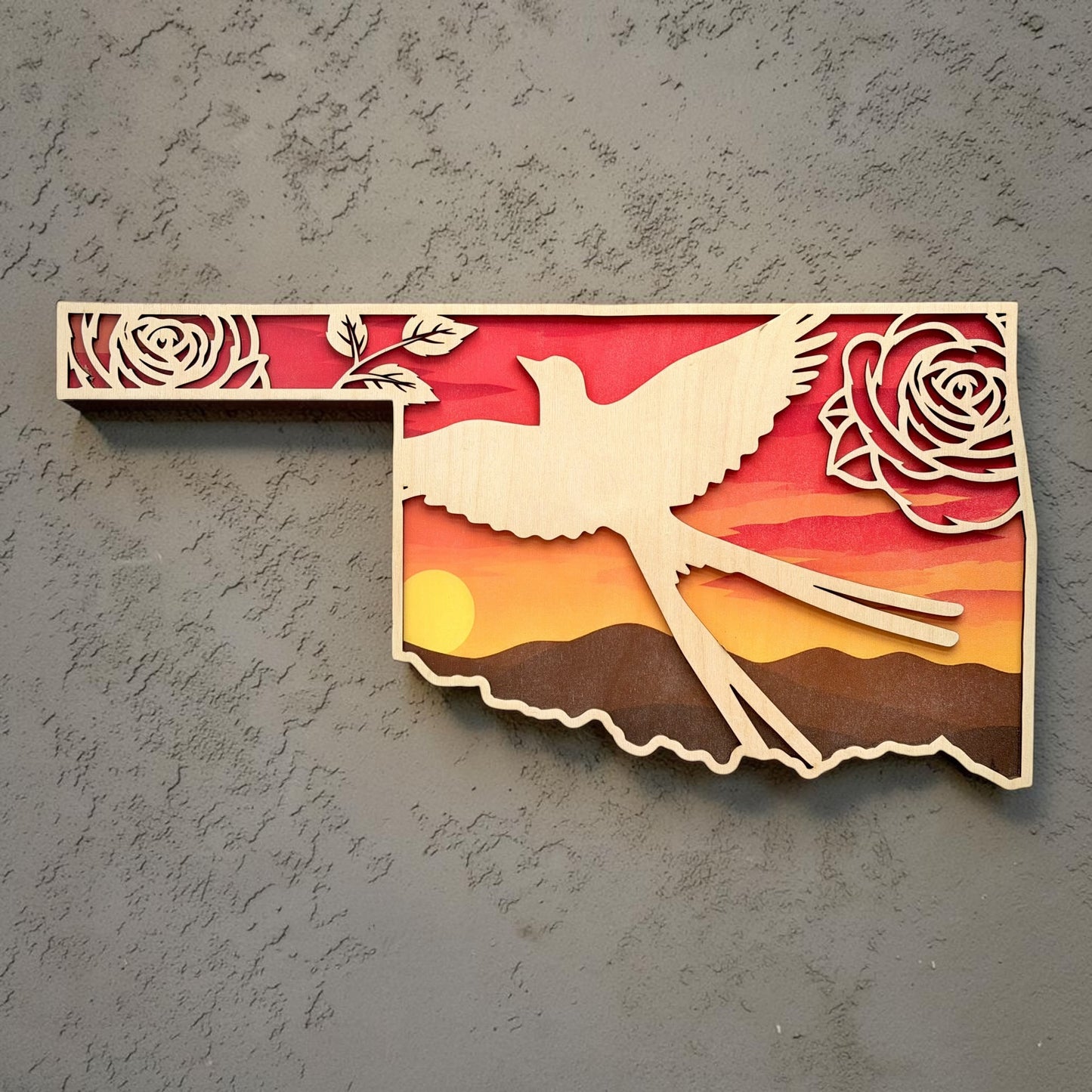 Oklahoma state shaped wall decor featuring a Scissortail Flycatcher silhouette, Oklahoma roses, and layered sunset prairie landscape displayed against textured gray wall background