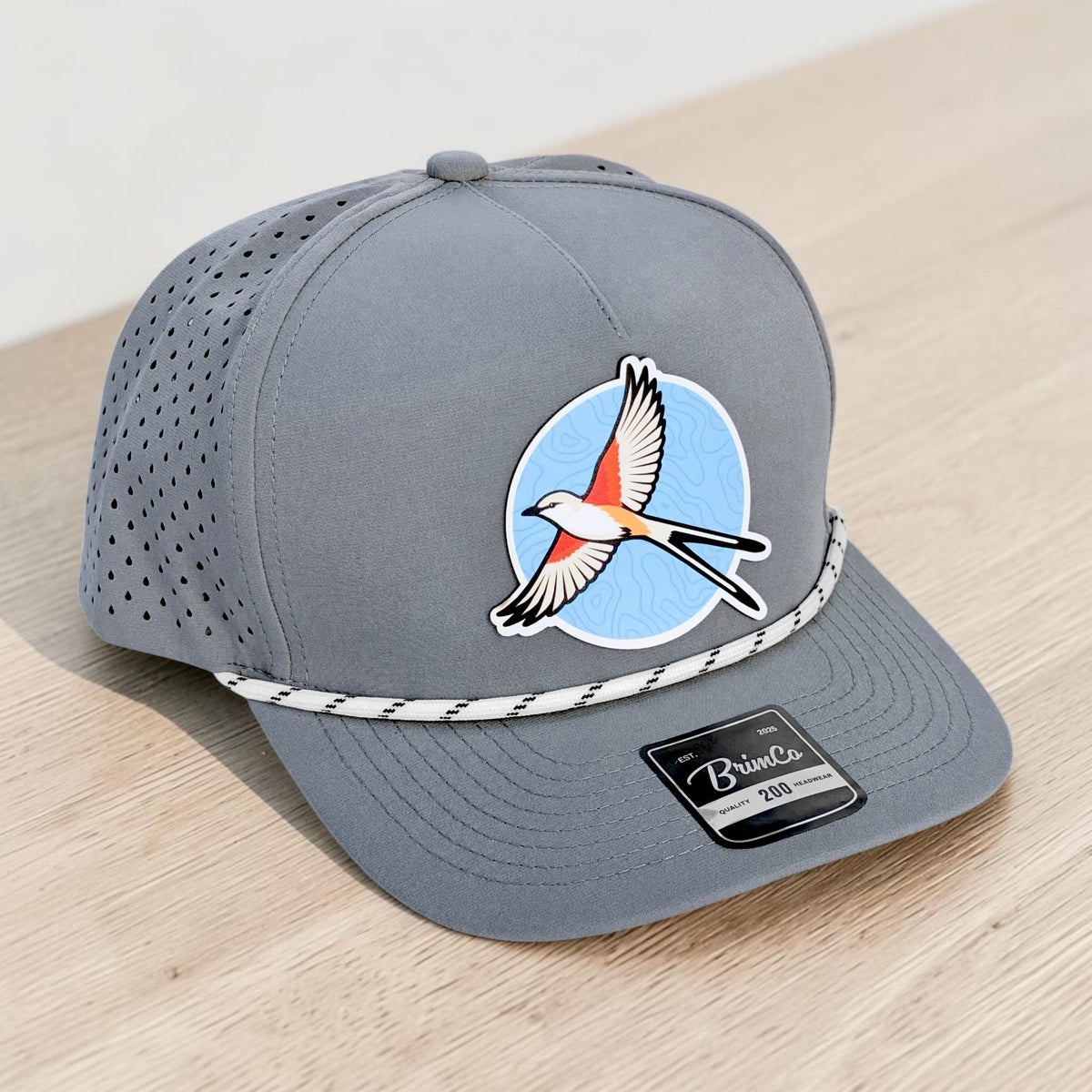 Angled view of gray Oklahoma scissortail performance golf hat showing colorful flycatcher patch, rope accent, precurved bill, and laser-perforated breathable side panels