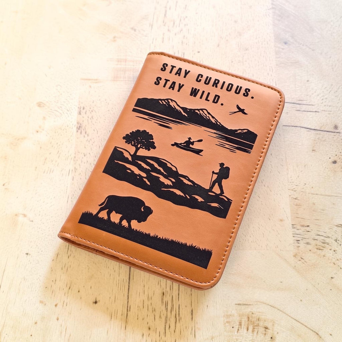 Angled view of engraved adventure passport wallet on wood table showing outdoor landscape artwork and stitched edges, travel wallet gift for hikers campers and road trip lovers
