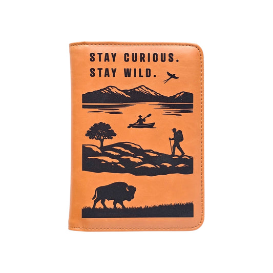 Brown leatherette passport wallet with Stay Curious Stay Wild text and engraved mountain lake hiking kayaking bird and bison artwork shown front view, adventure travel passport cover gift for travelers in the USA