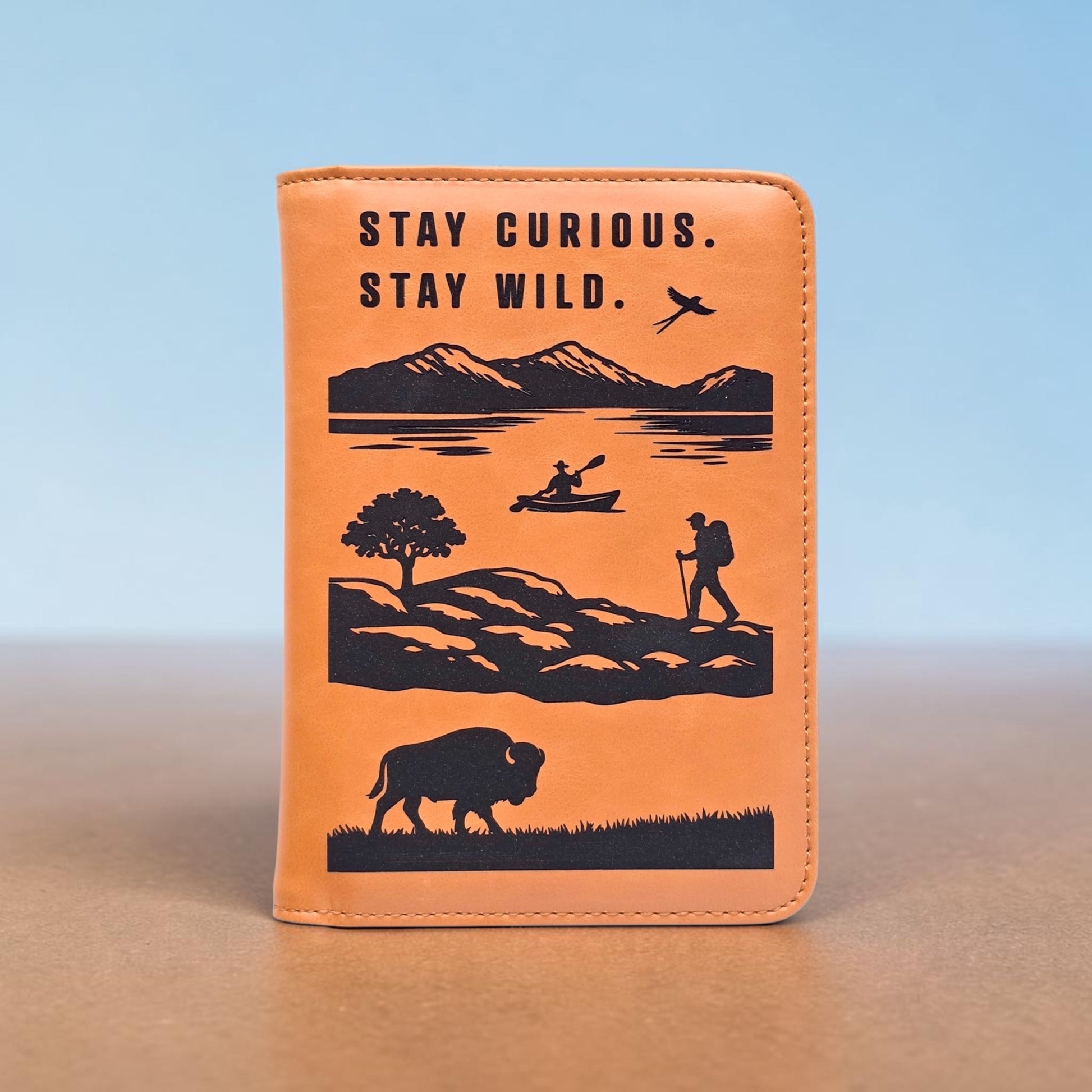 Front studio image of brown passport wallet with engraved mountains lake hiker kayaker bird and bison design, slim passport holder for travel organization
