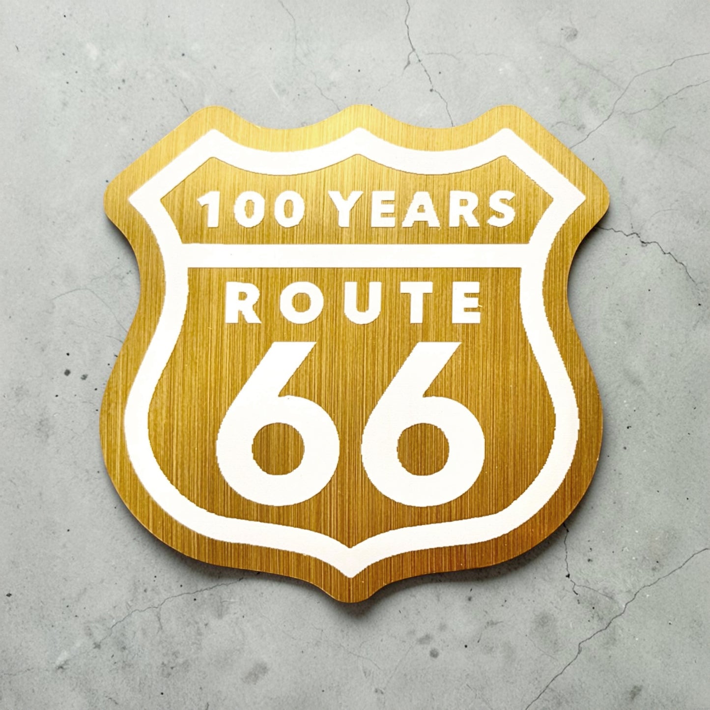 Route 66 100 years brushed gold vinyl sticker displayed on concrete background showing retro highway shield design used as a Route 66 souvenir decal or travel keepsake