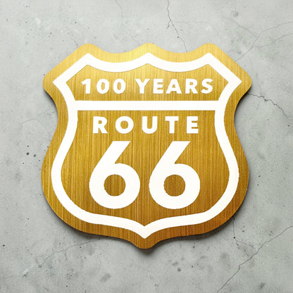 Route 66 100 years brushed gold vinyl sticker displayed on concrete background showing retro highway shield design used as a Route 66 souvenir decal or travel keepsake