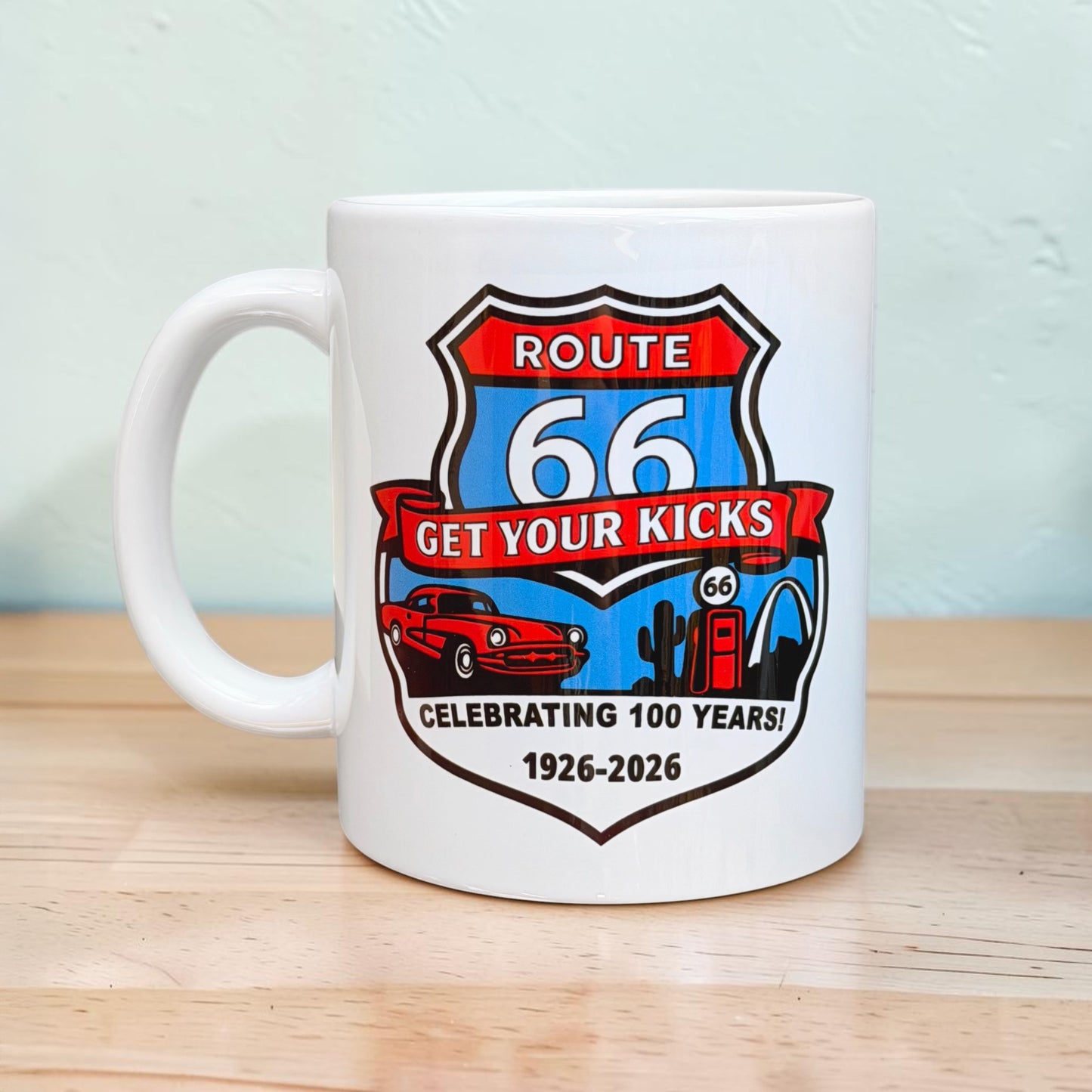 Route 66 Centennial ceramic coffee mug displayed on wood table featuring colorful highway shield artwork and nostalgic Americana road trip design for Route 66 travelers and souvenir collectors