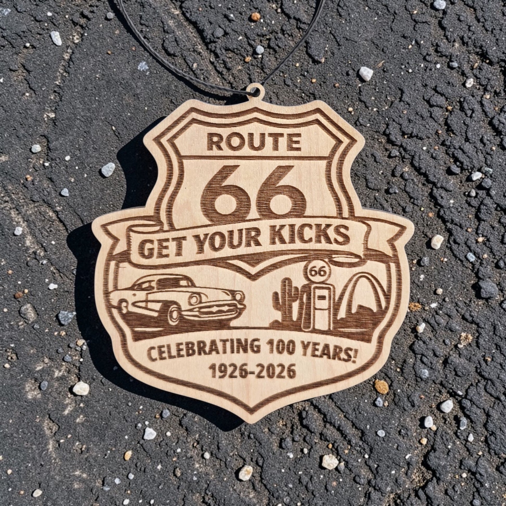 Route 66 Centennial wood ornament placed on asphalt road surface, engraved highway shield souvenir celebrating 1926 to 2026 travel history