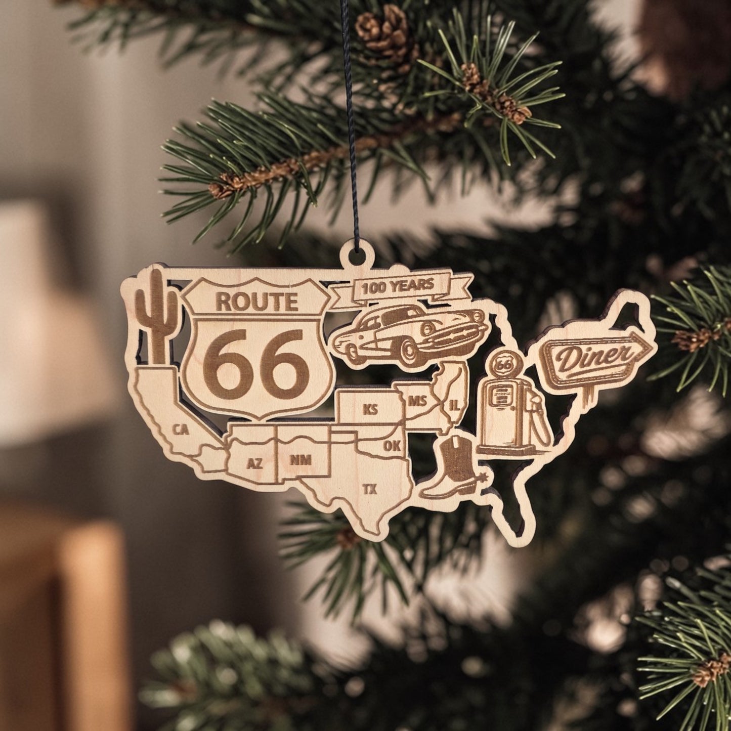 Route 66 centennial wooden ornament hanging on evergreen Christmas tree branch showing USA map design with classic road trip icons celebrating the historic Route 66 highway