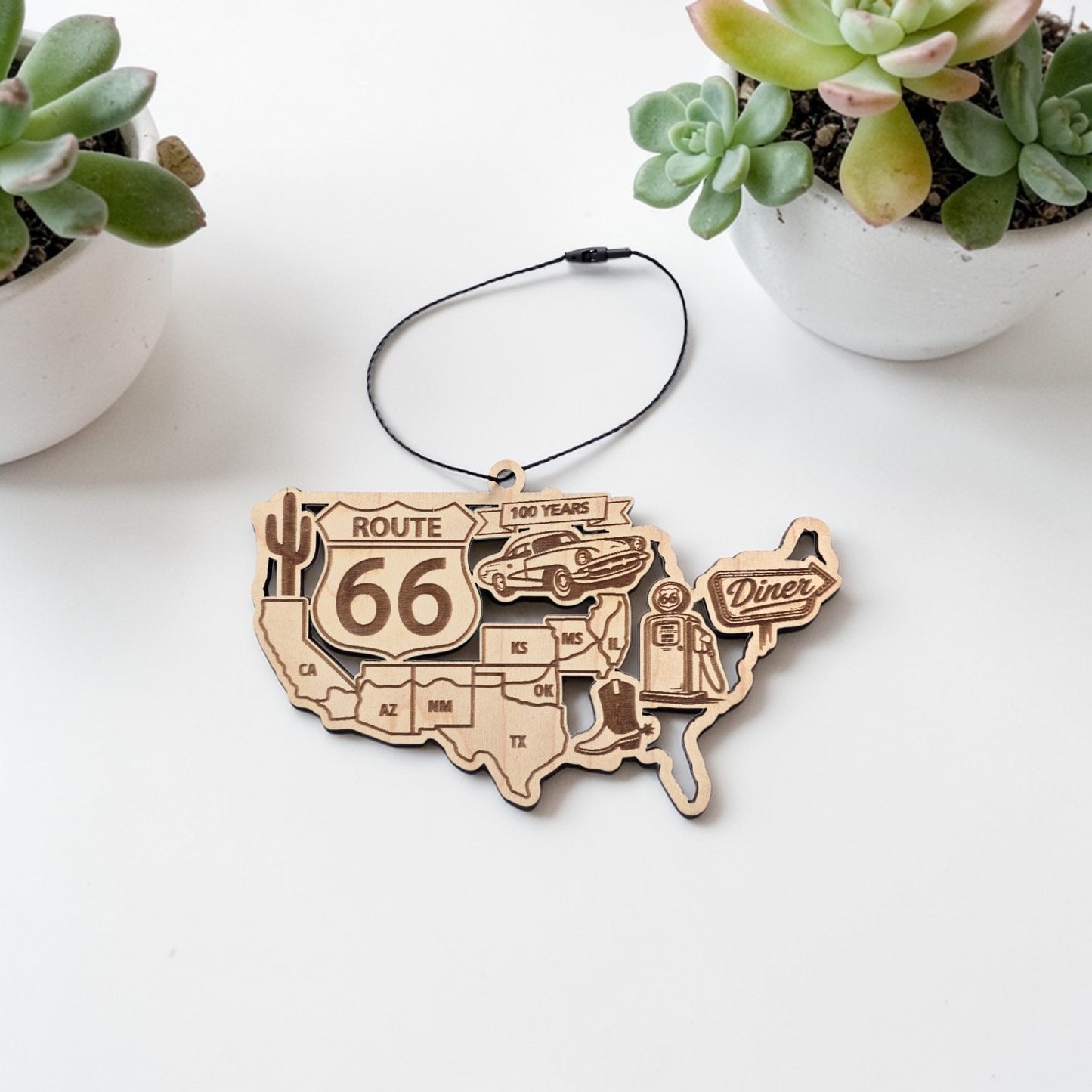 laser cut Route 66 centennial wooden ornament shaped like the United States map showing Route 66 states and vintage road trip icons including diner sign, gas pump, cactus and classic car