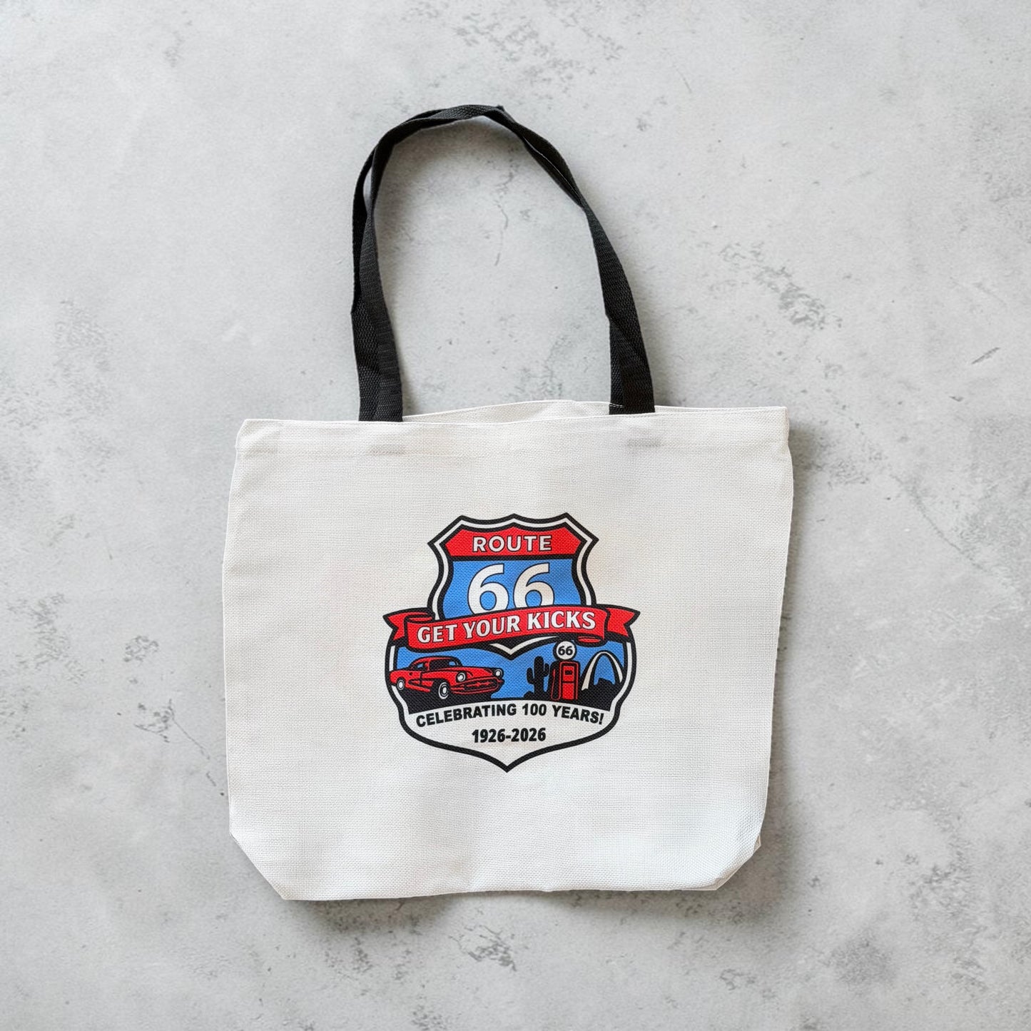 Flat lay view of Route 66 commemorative tote bag with vintage inspired Route 66 shield artwork, retro car, gas pump, and Americana travel icons on a white canvas style souvenir tote