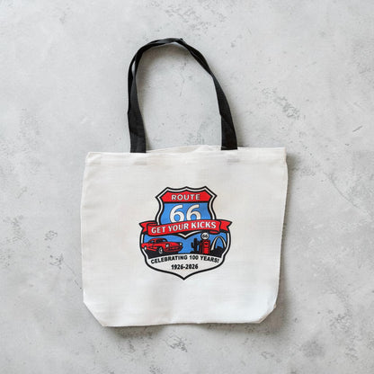 Flat lay view of Route 66 commemorative tote bag with vintage inspired Route 66 shield artwork, retro car, gas pump, and Americana travel icons on a white canvas style souvenir tote