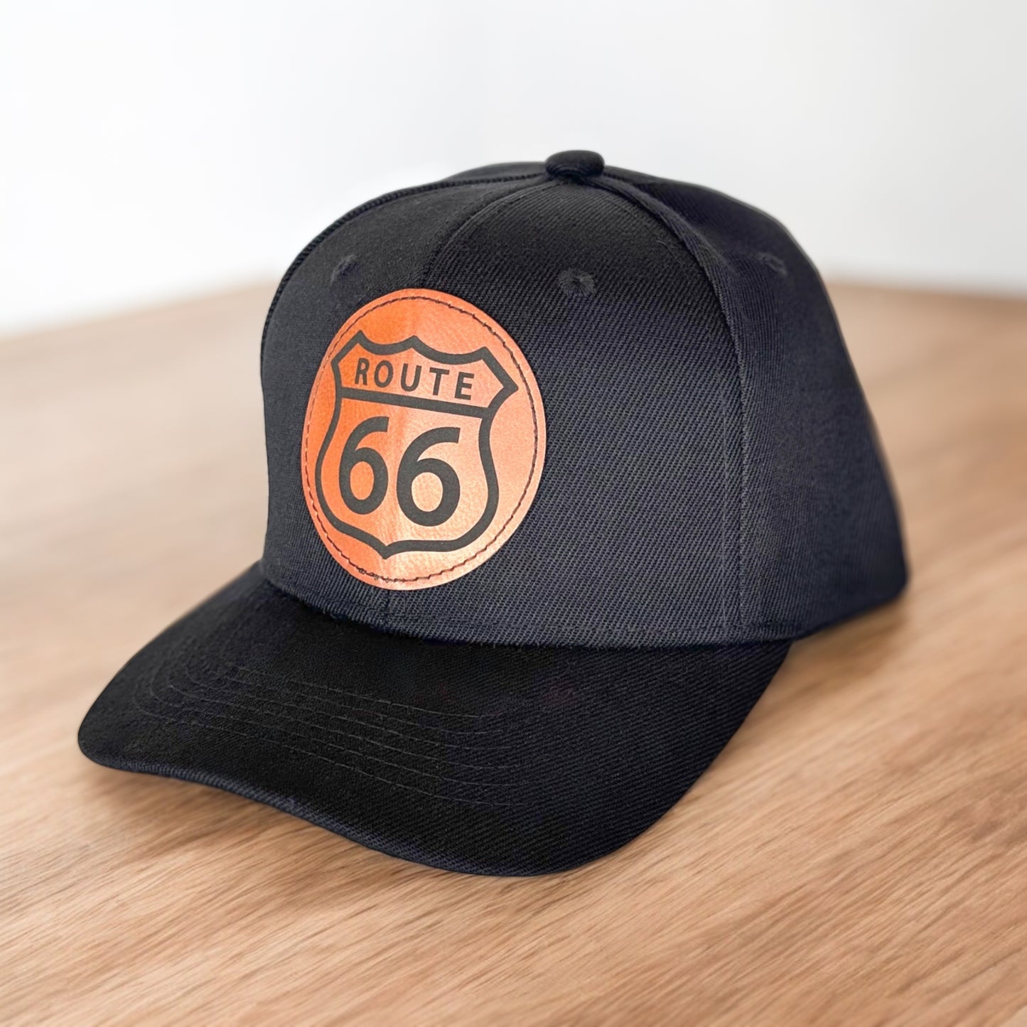 Angled view of black Route 66 leather patch hat on wooden surface, Oklahoma Route 66 trucker style cap