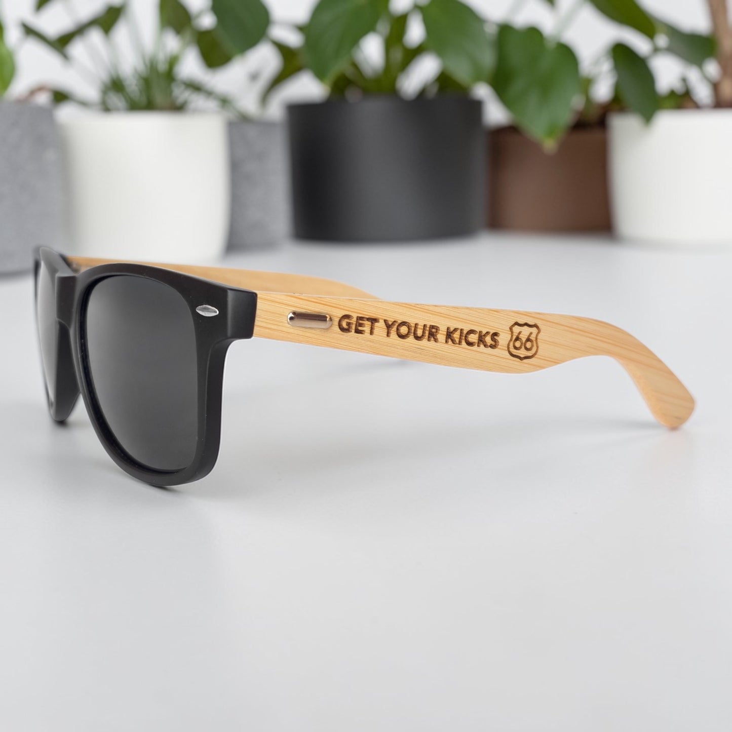 Close view of black sunglasses with engraved bamboo wood arms reading Get Your Kicks 66, Route 66 souvenir sunglasses displayed indoors with plants