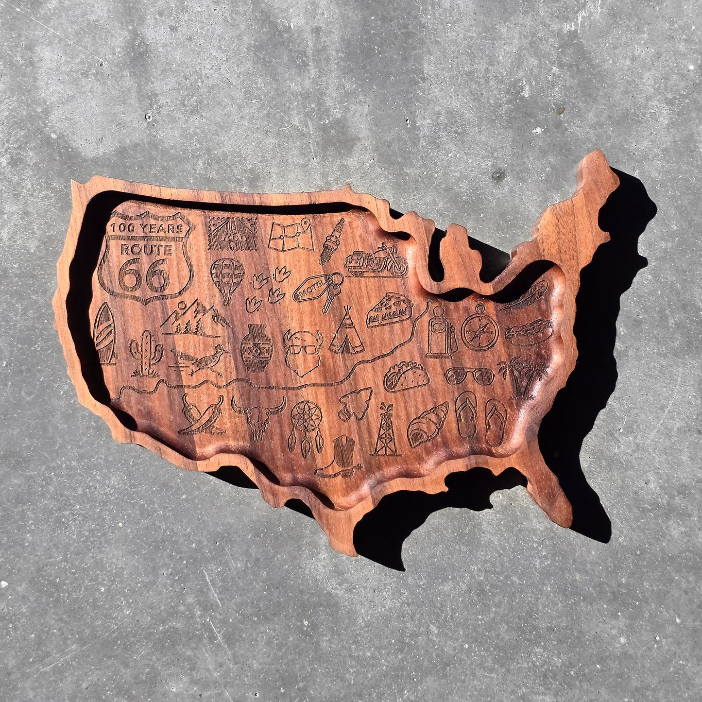 Hand carved USA shaped wooden tray displayed outdoors in sunlight featuring engraved Route 66 Centennial inspired travel icons and rustic Americana road trip details