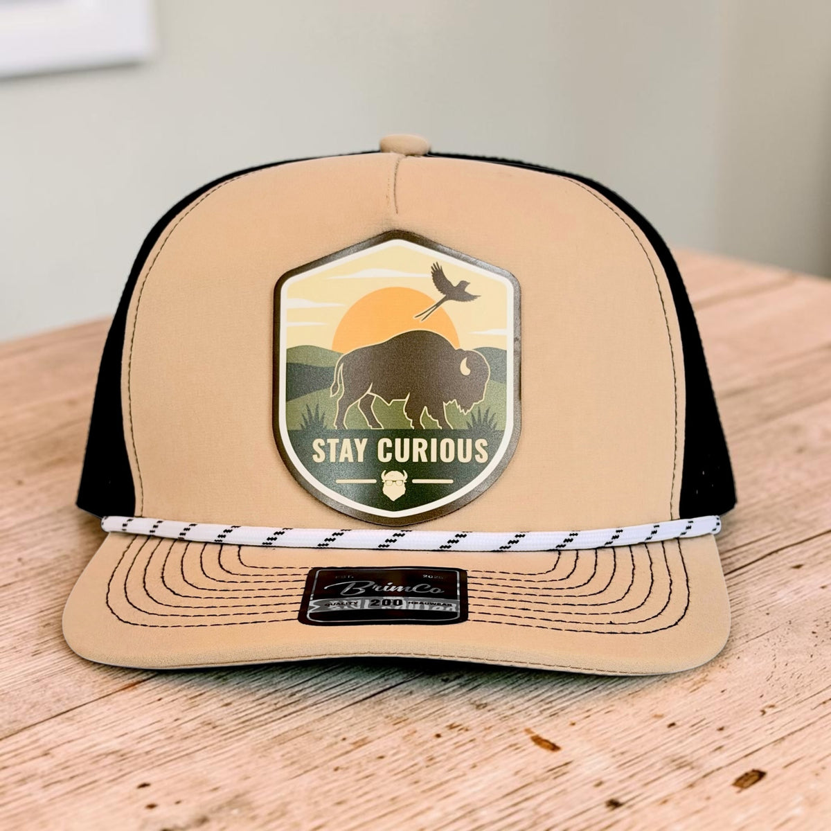 Tan and black performance rope golf hat displayed on a wood surface with colorful Stay Curious bison and Oklahoma scissortail patch on the structured front panel