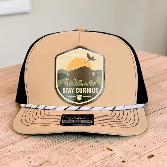 Tan and black performance rope golf hat displayed on a wood surface with colorful Stay Curious bison and Oklahoma scissortail patch on the structured front panel
