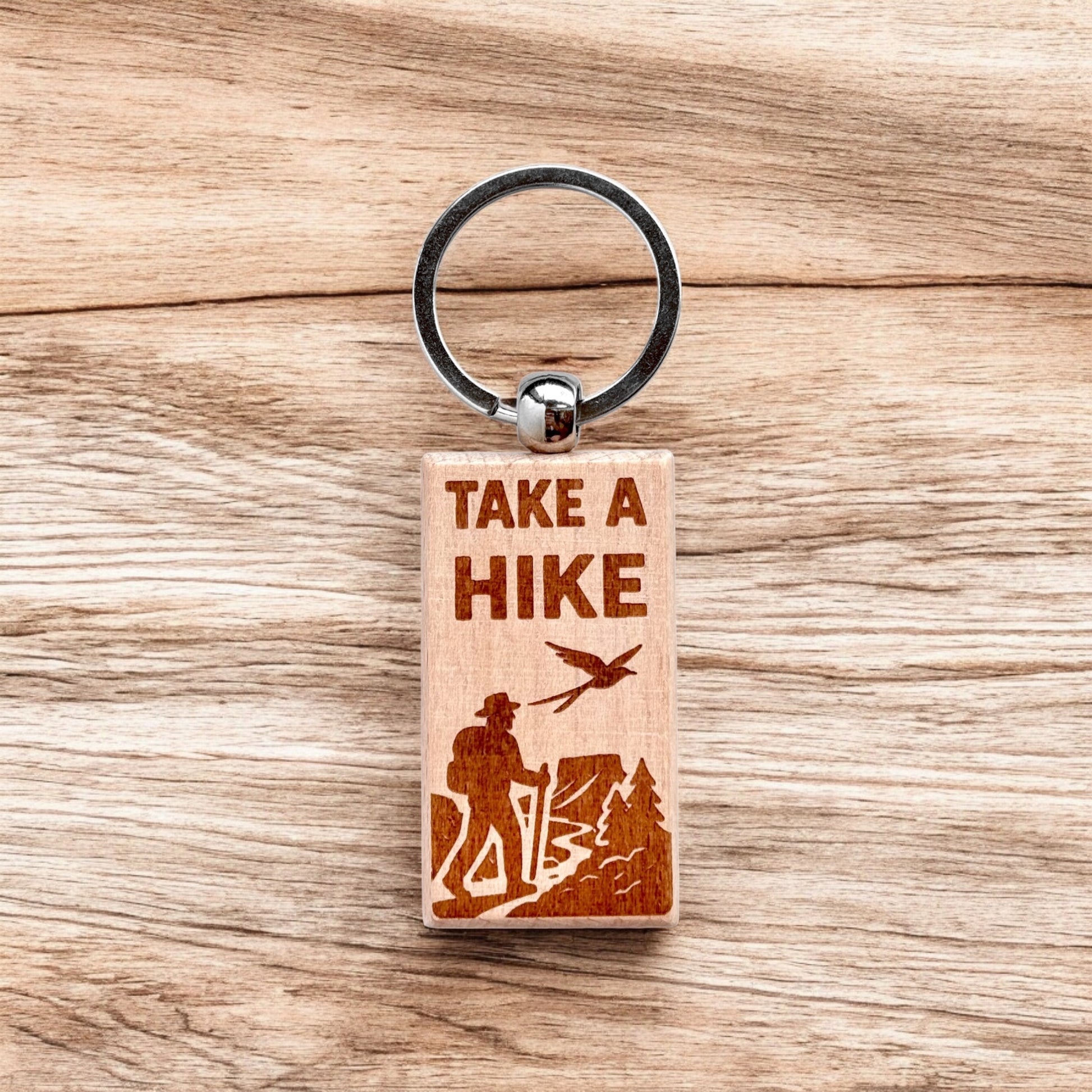 Laser engraved take a hike wooden keychain resting on wood surface, outdoor hiking themed wood keychain for camping gift, travel souvenir, or nature lover